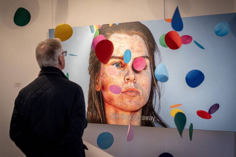 A man looking at the Here, There and Them Exhibition in Flowerfield Arts Centre. The painting is of a woman surrounded by falling coloured circles with the words “Still, they get away…”