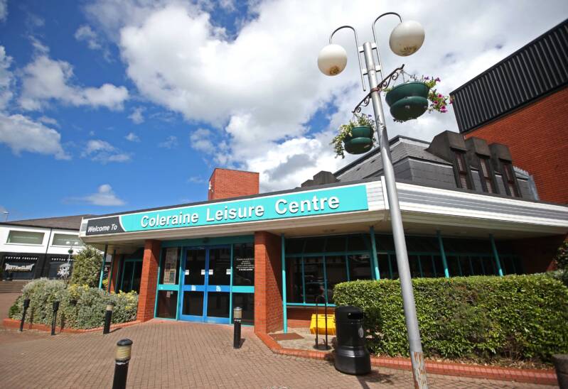 Image shows the entrance to Coleraine Leisure Centre, with hanging baskets and plants and a lamppost outside.