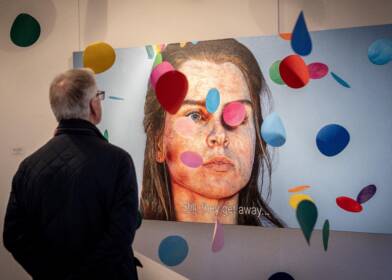 A man looking at the Here, There and Them Exhibition in Flowerfield Arts Centre. The painting is of a woman surrounded by falling coloured circles with the words “Still, they get away…”